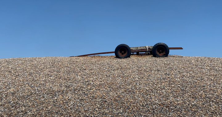 shingle beach, rusted trailer with one flat wheel with a bright blue sky