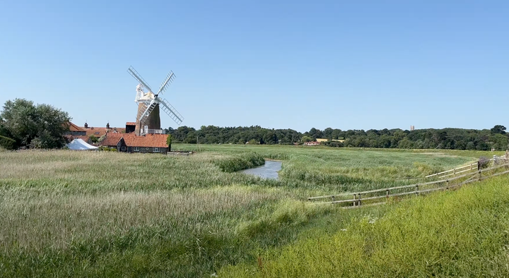 photograph of a windmill with white sails with a reed beds and a river in the foreground and trees on the horizon